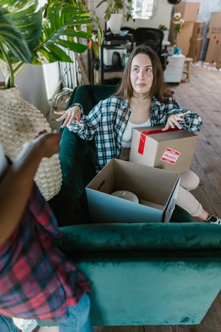 A person holding a cardboard box labeled 'BOOKS' is inside a room preparing for a house removal, with several other boxes stacked on a table in front of a woman who is seated on a bed. The woman, with