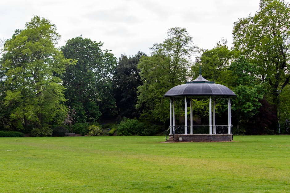 A small, open-sided pavilion with a dark, domed roof and white support columns is situated on a brick platform in the middle of a well-maintained grassy area within Dulwich Park. Surrounding the pavil