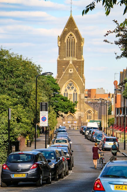 A street scene during daytime showing a row of parked cars along the curb, with a woman and a person on a bicycle moving along the pavement. In the background, there is a large, historic stone church with a tall, pointed steeple and Gothic-style arched windows, partially obscured by green leafy trees lining the street. The scene features various street lamps, traffic signs, and buildings on both sides, with clear weather and a blue sky. This image relates to house removals and moving services, highlighting urban logistics, loading and unloading areas, and the importance of careful planning in home relocation processes, as provided by Removals Dulwich.