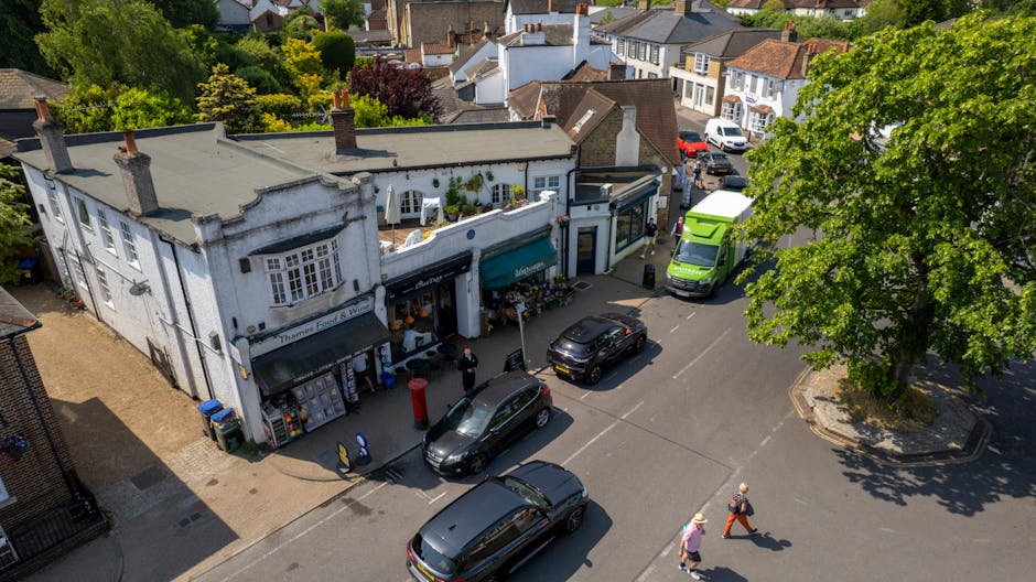 Aerial view of a busy street in Dulwich Village with a white two-storey building featuring multiple shopfronts, including a post box and large windows displaying products. Several vehicles, such as a green van and multiple cars, are parked along the road. Two pedestrians walk across the street, and a large leafy tree provides shade on the right side. The scene captures both retail activity and the surrounding residential and commercial buildings, highlighting the typical environment for home relocation and furniture transport services offered by Removals Dulwich.