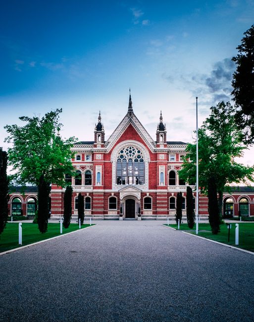 A grand, historic building constructed with red brick and white stone accents, featuring large arched windows, decorative gables, and multiple spires on the roof. The structure is set against a partly cloudy sky during daylight, with two tall trees on either side of a wide paved pathway leading up to the entrance. In front of the building, there is a neatly maintained lawn with small white bollards along the edge of the pathway. The scene appears calm and well-kept, suitable for hosting events or institutional use. This architecture is representative of a classic civic or community building, with no interior or moving activity visible in the image, aligning with the context of professional house removals and relocation services by Removals Dulwich.