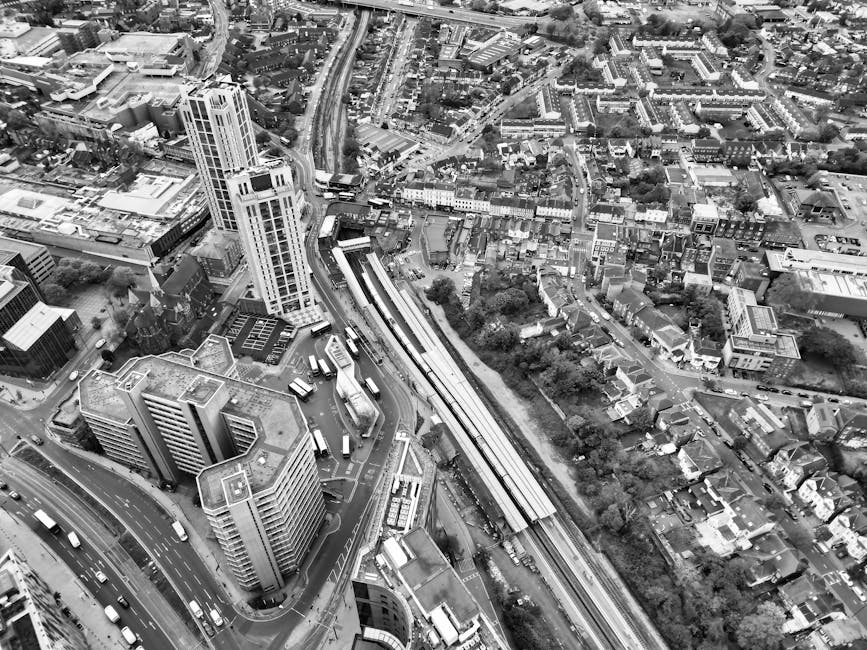 A black and white aerial photograph of a densely developed urban area showing multiple high-rise and mid-rise buildings, city streets with vehicles, and railway tracks running through the middle. The scene includes a mix of commercial and residential structures, with some greenery and trees interspersed among the buildings. The railway tracks are positioned centrally, with a partially visible train station platform adjacent to them, and an overpass crossing above the tracks. The image captures the busy, layered nature of city infrastructure, with roads, buses, and cars visible at street level. This urban landscape image is relevant to house removals and relocation services such as those offered by Removals Dulwich, illustrating the typical environment for home relocation and furniture transport within a cityscape.